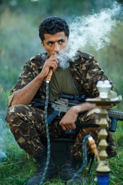 Un homme en camouflage avec un fusil fume un narguilé en plein air — Photo