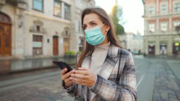Пандемия коронавируса Ковид-19. Женщина в защитной медицинской маске идет по улице и пользуется смартфоном — стоковое видео