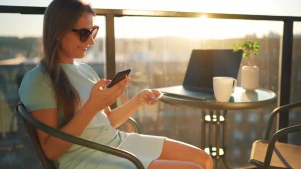 Mujer sentada en el balcón contra el telón de fondo del sol poniente y hace una compra en línea con una tarjeta de crédito y un teléfono inteligente — Vídeo de stock