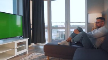 Evdeki adam kanepede uzanıp yeşil ekran TV izliyor.