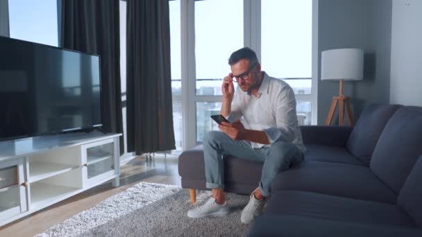 Hombre con gafas sentado en el sofá en una habitación acogedora y el uso de teléfono inteligente para navegar por Internet o charlar en mensajeros — Vídeos de Stock