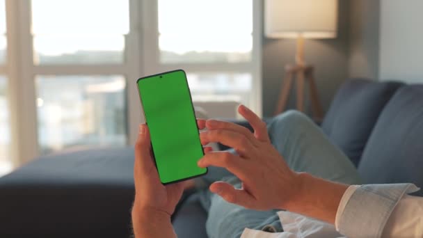 Hombre en casa acostado en un sofá y usando un teléfono inteligente con pantalla de maqueta verde en modo vertical. Chico navegando por Internet, viendo contenido, videos, blogs. POV. — Vídeos de Stock