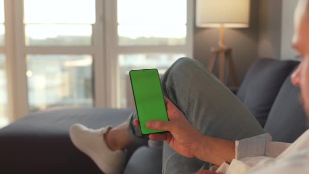自宅の男はソファに横になり、垂直モードで緑色のモックアップ画面を持つスマートフォンを使用しています。男は、コンテンツ、ビデオ、ブログを見て、インターネットを閲覧. — ストック動画