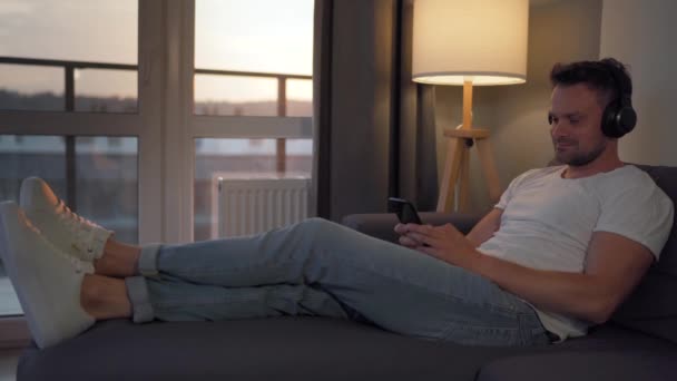 Hombre con auriculares se encuentra en el sofá, escucha música y utiliza un teléfono inteligente para comunicarse en mensajeros instantáneos o redes sociales — Vídeo de stock