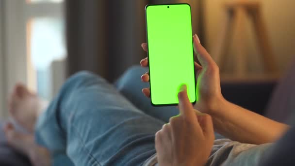 Mujer en casa acostada en un sofá y usando un smartphone con pantalla verde en modo vertical. Chica navegando por Internet, viendo contenido, videos, blogs. POV. — Vídeo de stock