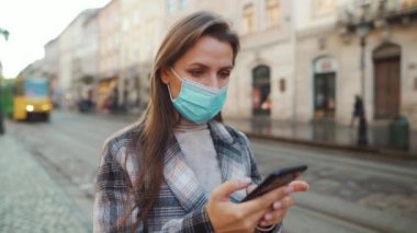 Covid-19 koronavirüsünün pandemik koruması. Koruyucu tıbbi maskeli bir kadın sokakta dikiliyor ve akıllı telefon kullanıyor.