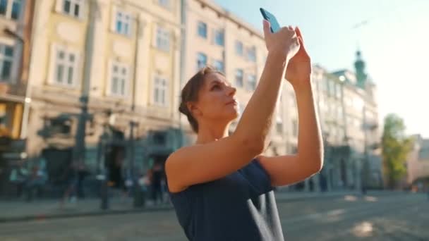 Mulher fica em uma rua velha e tira uma foto ou vídeo em um smartphone ao pôr do sol — Vídeo de Stock