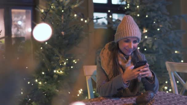Зимою жінка сидить у кафе на вулиці і розмовляє в соціальних мережах, використовуючи смартфон з різдвяними прикрасами на задньому плані. — стокове відео