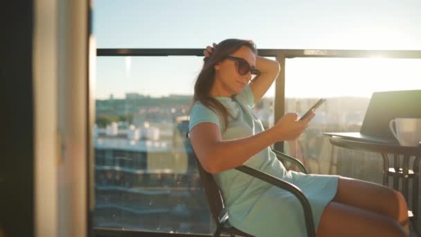 Wanita menggunakan smartphone di balkon terhadap latar belakang matahari terbenam. Kantor luar rumah. Konsep kerja jarak jauh — Stok Video