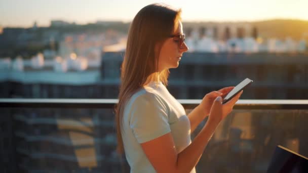 Žena používající smartphone na balkóně na pozadí západu slunce. Ona chatuje s někým on-line a posílá fotku západu slunce. — Stock video