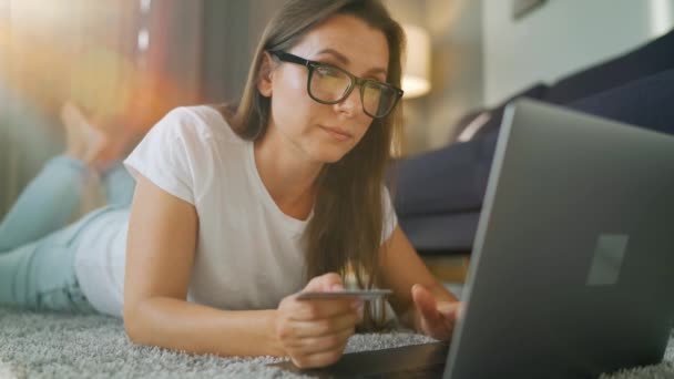 Mujer con gafas está tirado en el suelo y hace una compra en línea con una tarjeta de crédito y un ordenador portátil. Compras en línea, tecnología de estilo de vida — Vídeo de stock
