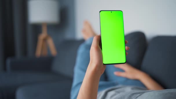 Mujer en casa acostada en un sofá y usando un smartphone con pantalla verde en modo vertical. Chica navegando por Internet, viendo contenido, videos, blogs. POV. — Vídeos de Stock