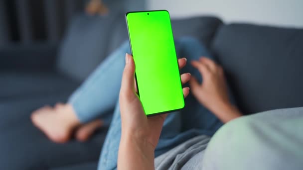 Mujer en casa acostada en un sofá y usando un smartphone con pantalla verde en modo vertical. Chica navegando por Internet, viendo contenido, videos, blogs. POV. — Vídeos de Stock