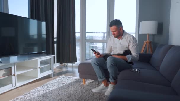 Muž s brýlemi sedí na gauči v útulném pokoji a používá smartphone. Hladí černou kočku, která mu leží na klíně a bere si selfie — Stock video