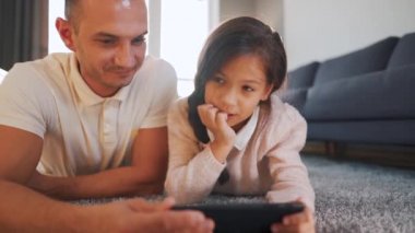 Mutlu gülümseyen baba ve kız birlikte vakit geçiriyorlar ve evde oturma odasında yerde yatarken aile eğlencesi için tablet kullanıyorlar..