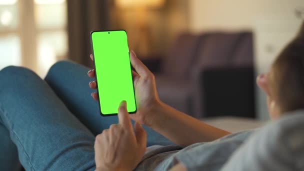 Frau zu Hause auf dem Sofa liegend und Smartphone mit grünem Bildschirm im vertikalen Modus. Mädchen surfen im Internet, schauen Inhalte, Videos, Blogs. — Stockvideo