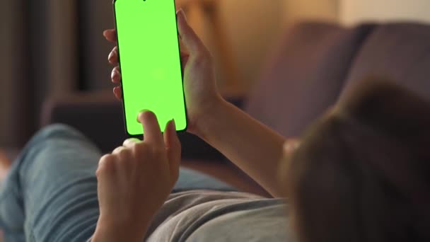 Vrouw thuis liggend op een bank en met behulp van smartphone met groene mock-up scherm in verticale modus. Meisje surfen op internet, kijken naar inhoud, video 's, blogs. — Stockvideo