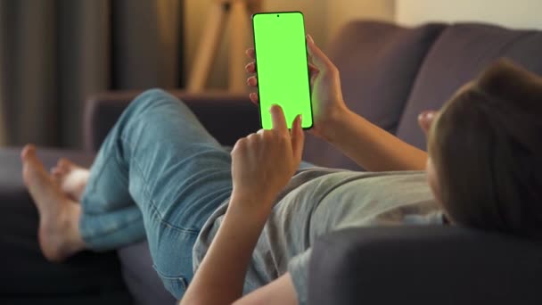 Mujer en casa acostada en un sofá y usando un smartphone con pantalla verde en modo vertical. Chica navegando por Internet, viendo contenido, videos, blogs. — Vídeo de stock