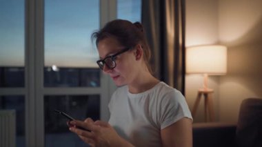 Gözlüklü bir kadın rahat bir odada kanepede oturuyor ve akşamları internette sörf yapmak için akıllı telefon kullanıyor. Rahatlama ve yaşam tarzı teknolojisi.