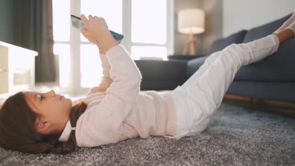 Niña usando tableta digital acostada boca arriba en el suelo en una habitación acogedora — Vídeo de stock