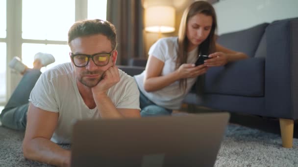 カップルは居心地の良い部屋で一緒に時間を過ごします:男はカーペットの上にあり、インターネットをサーフィンするためにスマートフォンを使用してノートパソコン、女性を使用しています。リモートワークの概念 — ストック動画
