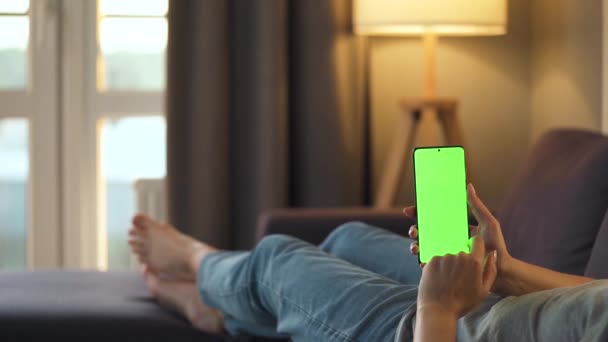 Donna a casa sdraiata su un divano e utilizzando smartphone con schermo mock-up verde in modalità verticale. Ragazza che naviga in Internet, guarda contenuti, video, blog. Luogo di nascita:. — Video Stock