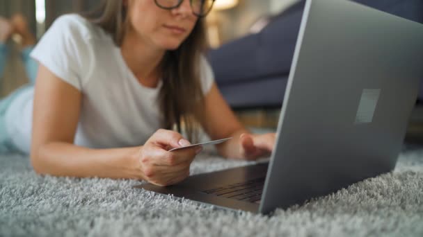 Kobieta w okularach leży na podłodze i dokonuje zakupu online za pomocą karty kredytowej i laptopa. Zakupy online, technologia stylu życia — Wideo stockowe