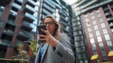 Resmi olarak giyinmiş bir kadın elinde kahveyle bir iş bölgesinde duruyor ve akıllı telefon kullanıyor.