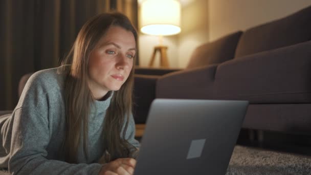 Kobieta leży na dywanie i wieczorem pracuje nad laptopem. Pojęcie pracy zdalnej. — Wideo stockowe