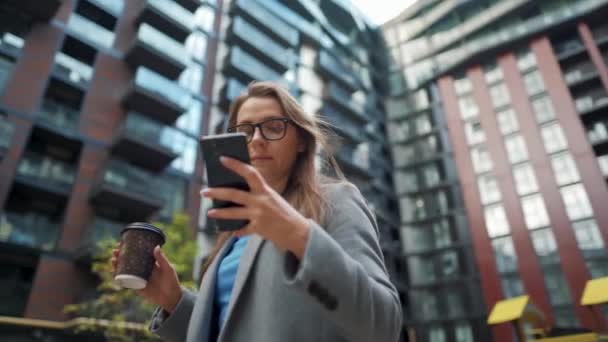 Formeel geklede vrouw staat in een zakenwijk met koffie in de hand en gebruikt een smartphone — Stockvideo