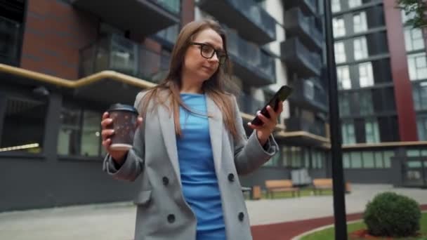 Mujer vestida formalmente camina por la calle en un distrito de negocios con café en la mano y utiliza un teléfono inteligente — Vídeo de stock