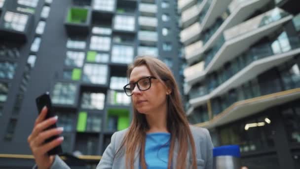Formeel geklede vrouw loopt door de straat in een zakenwijk met thermoskan in de hand en maakt gebruik van een smartphone — Stockvideo