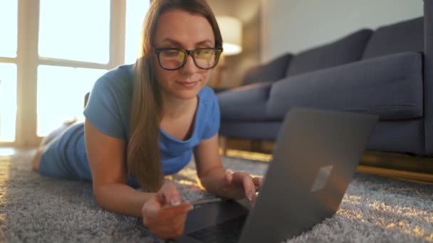 Mulher com óculos está deitada no chão e faz uma compra on-line usando um cartão de crédito e laptop. Compras online, tecnologia de estilo de vida — Vídeo de Stock