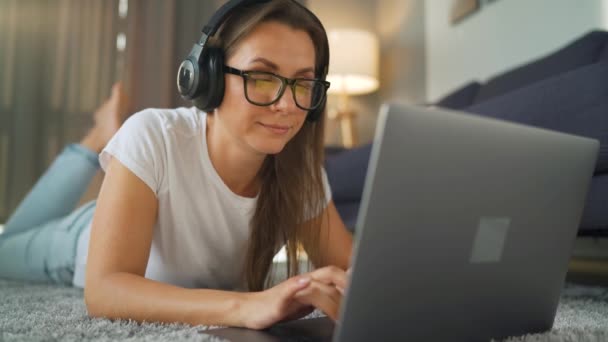 Damen med briller ligger på gulvet, jobber med en bærbar datamaskin og hører på musikk på hodetelefoner. Fjernarbeidsbegrep. – stockvideo