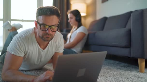 Pareja pasa tiempo juntos en una habitación acogedora: el hombre se acuesta en una alfombra y utiliza un ordenador portátil, mujer con auriculares escucha música y utiliza el teléfono inteligente para navegar por Internet. Concepto de trabajo remoto — Vídeo de stock