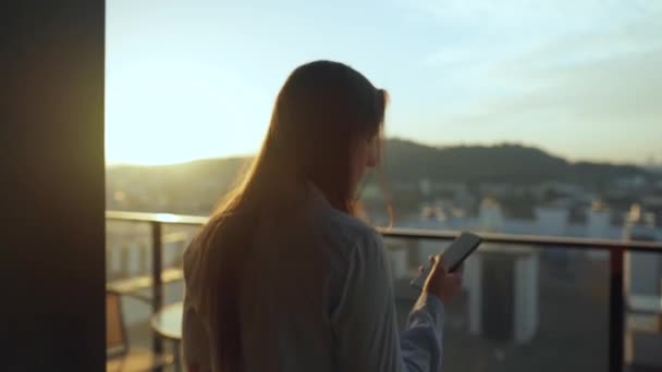 Kvinnan börjar sin dag med en kopp te eller kaffe och kollar e-post i sin smartphone på balkongen i gryningen. — Stockvideo