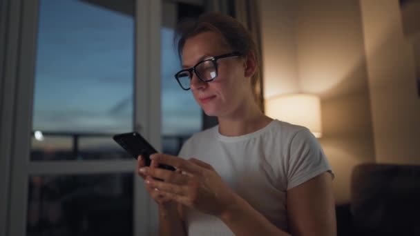 Wanita dengan kacamata duduk di sofa di kamar yang nyaman dan menggunakan smartphone untuk surfing internet di malam hari. Teknologi relaks dan gaya hidup. — Stok Video