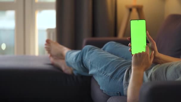 Woman at home lying on a sofa and using smartphone with green mock-up screen in vertical mode. Girl browsing Internet, watching content, videos, blogs. POV. — Stock Video