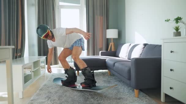 Vídeo divertido. El hombre en pantalones cortos y una camiseta representa el snowboard en una alfombra en una habitación acogedora. Esperando un invierno nevado — Vídeos de Stock