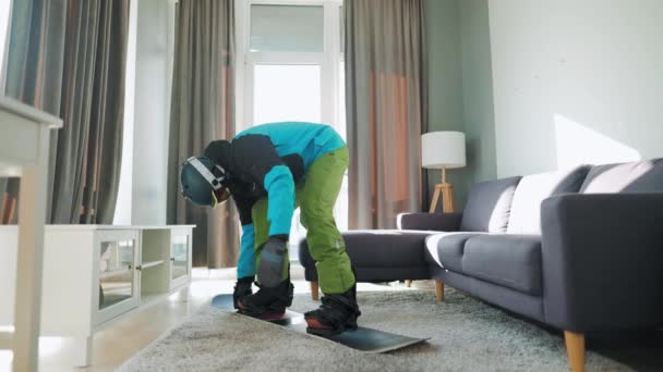 Vídeo divertido. Hombre vestido de snowboarder monta una tabla de snowboard en una alfombra en una habitación acogedora. Esperando un invierno nevado — Vídeos de Stock