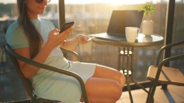 Batan güneşin arka planında balkonda oturan bir kadın kredi kartı ve akıllı telefon kullanarak online alışveriş yapıyor.