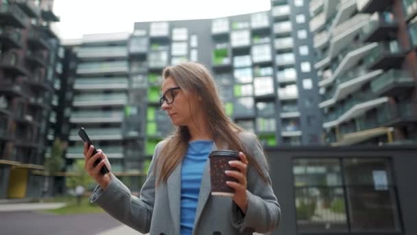 O femeie îmbrăcată formal stând într-un cartier de afaceri cu cafea în mână și folosind un smartphone. Camera se mișcă în jurul ei — Videoclip de stoc