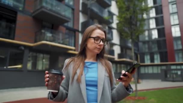 Donna formalmente vestita che cammina per strada in un quartiere degli affari con il caffè in mano e utilizzando uno smartphone — Video Stock