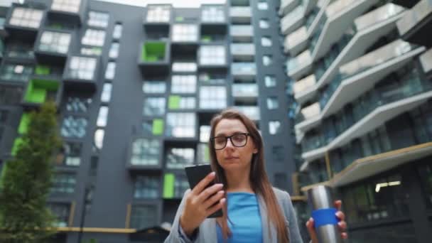 Formell gekleidete Frau läuft mit Thermobecher in der Hand und Smartphone durch ein Geschäftsviertel — Stockvideo