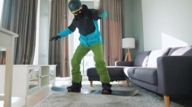 Eğlenceli video. Snowboard 'cu gibi giyinmiş bir adam rahat bir odada halının üstünde snowboard sürüyor. Karlı bir kış bekliyor