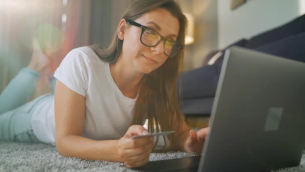 안경을 쓴 여성은 바닥에 누워 신용 카드와 노트북을 사용하여 온라인 구매를 한다. 온라인 쇼핑, 생활 방식 기술 — 비디오