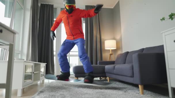 Vídeo divertido. Hombre vestido de snowboarder monta una tabla de snowboard en una alfombra en una habitación acogedora. Esperando un invierno nevado — Vídeos de Stock