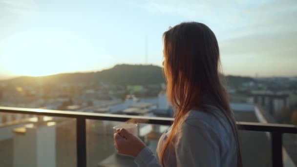 美しい都市の景色を眺めながら、美しい女性はコーヒーや紅茶を飲みながら、日の出にバルコニーで新鮮な冷気を呼吸するリラックスをお楽しみください。 — ストック動画