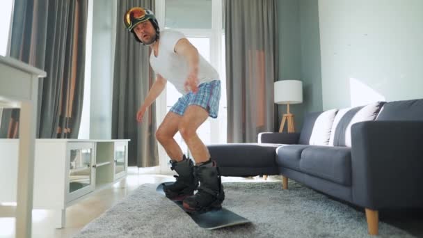 Lustiges Video. Ein Mann in kurzen Hosen und T-Shirt zeigt Snowboarder auf einem Teppich in einem gemütlichen Raum. Warten auf einen schneereichen Winter — Stockvideo