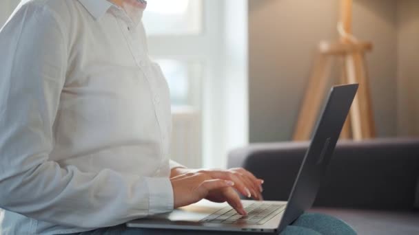 眼鏡をかけた女性が居心地の良い部屋のソファに座ってノートパソコンで作業しています。リモートワークの概念. — ストック動画
